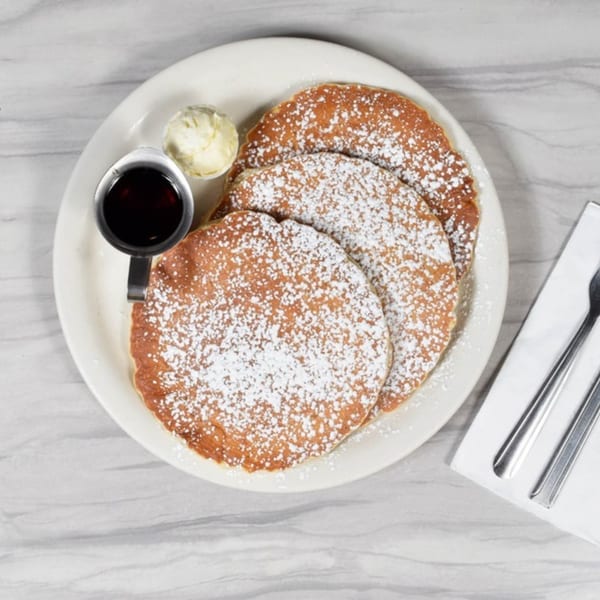 Three pancakes dusted with powdered sugar, with a side of butter and syrup, on a white plate.
