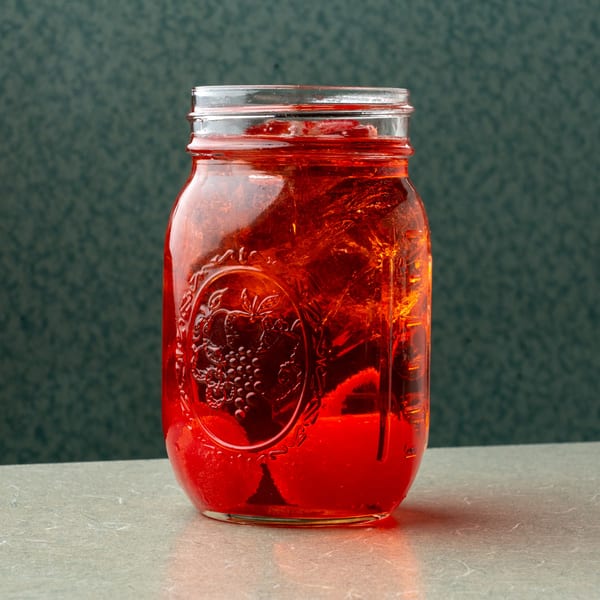 A mason jar filled with a vibrant red drink and ice, sitting on a light-colored surface.