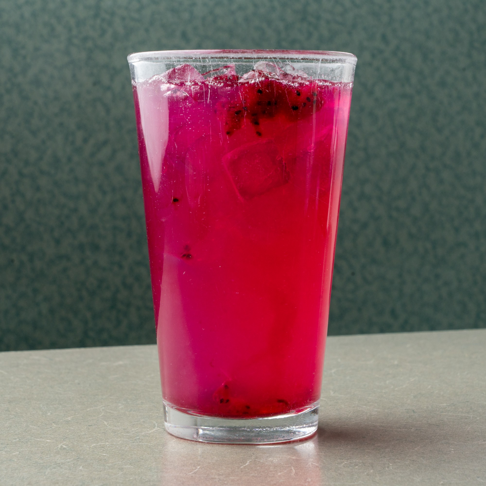 Vibrant pink dragon fruit drink with ice in a clear glass on a diner table.