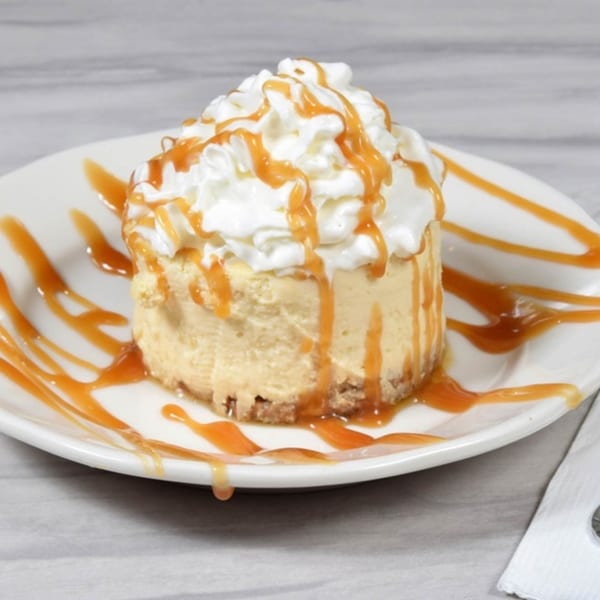 Mini cheesecake topped with whipped cream and drizzled with caramel sauce on a white plate.
