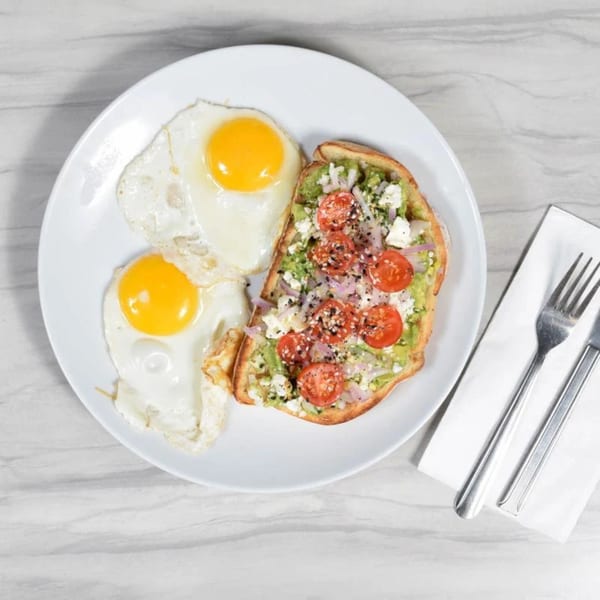 Two fried eggs and avocado toast with tomatoes, feta, and red onion on a white plate.